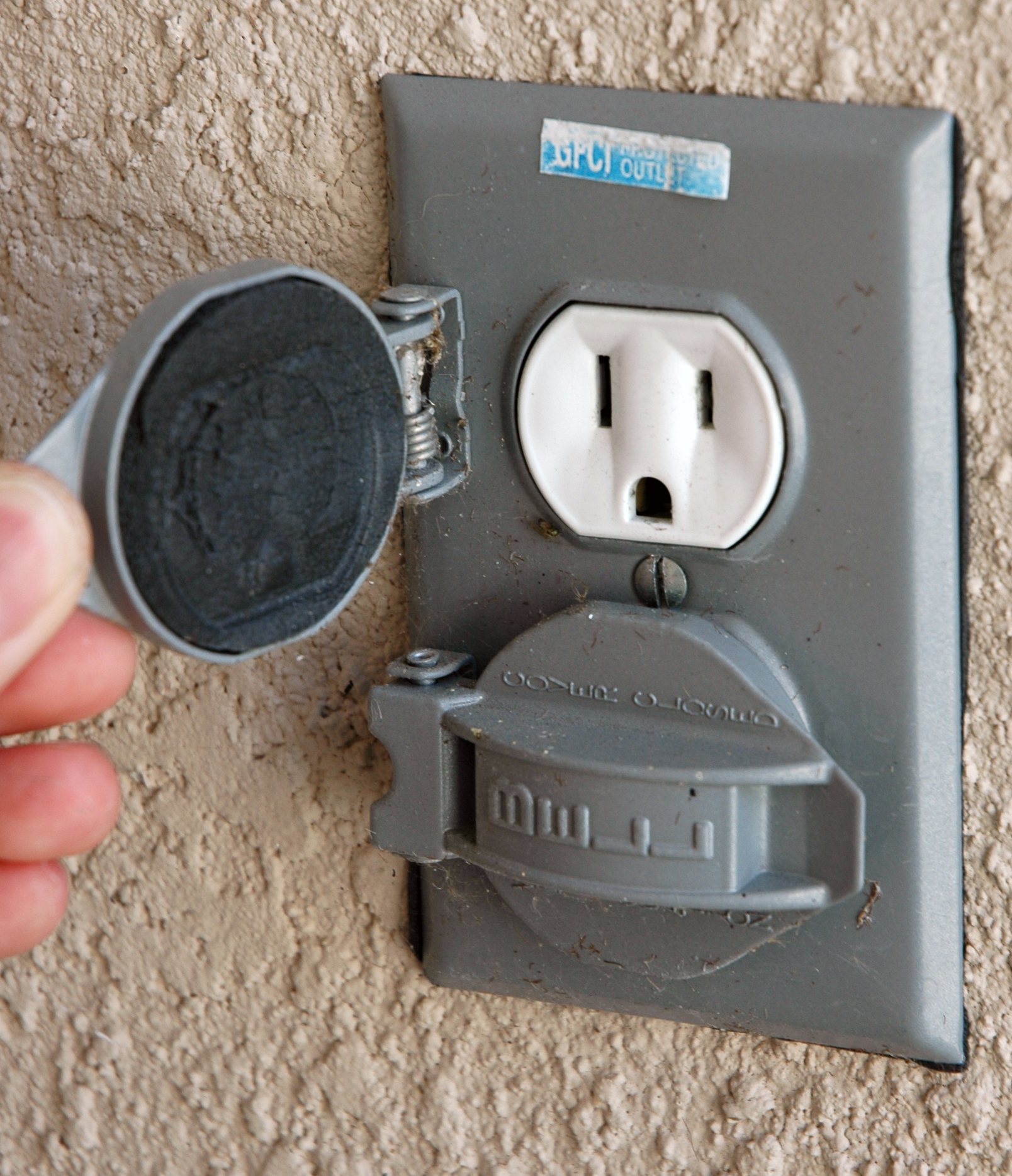 Outdoor U.S. electrical outlet with a hinged metal cover and GFCI-protected label.