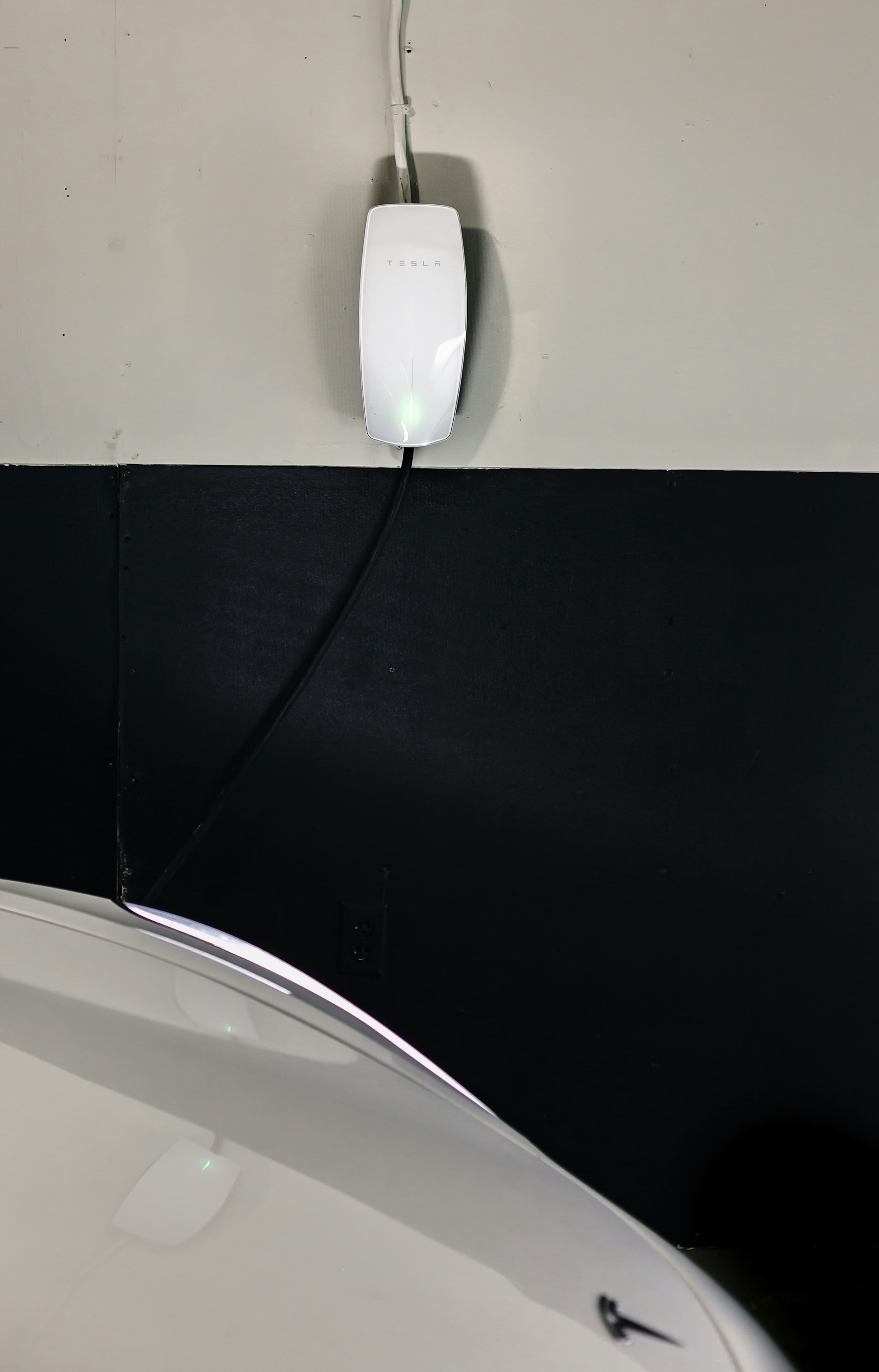 Wall-mounted electric vehicle charging unit and cable beside a white car in a garage.