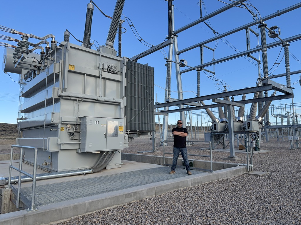 An electrician beside a large transformer and electrical equipment at an outdoor substation